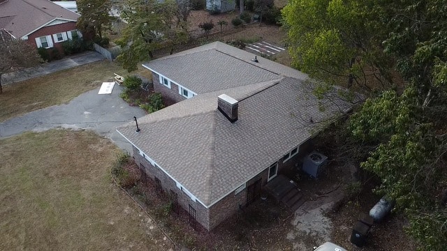 aerial view of a single-story brick house with a gray shingled roof, chimney, and surrounding yard.