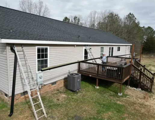 gutter-gard Backyard of a beige single-story house with a raised wooden deck; two workers on the deck hold a long wooden beam, with ladders leaning against the house and an AC unit nearby.