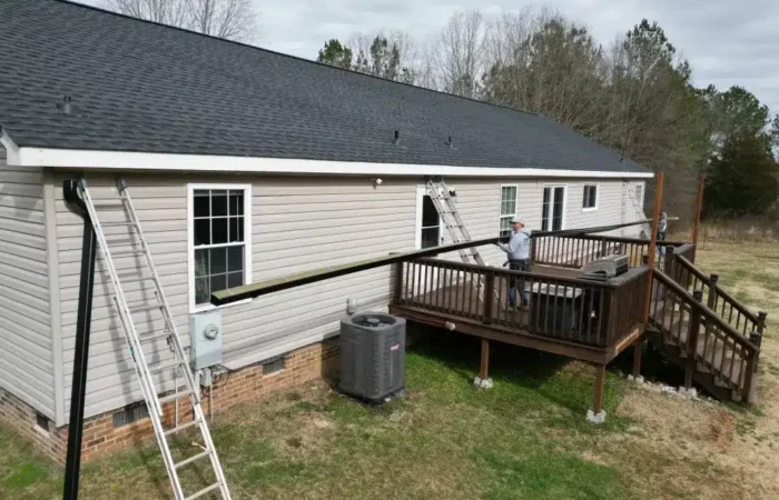 backyard of a beige single-story house with a raised wooden deck; two workers on the deck hold a long wooden beam, with ladders leaning against the house and an ac unit nearby.