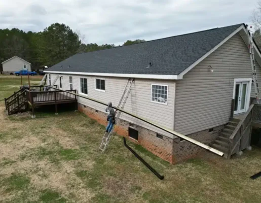 gutter-installation Beige single-story house with brick foundation; contractor on a ladder repairing the eaves near the deck. A long ladder rests on the ground beside the house, with another ladder on the roof edge and a wooden deck on the left.